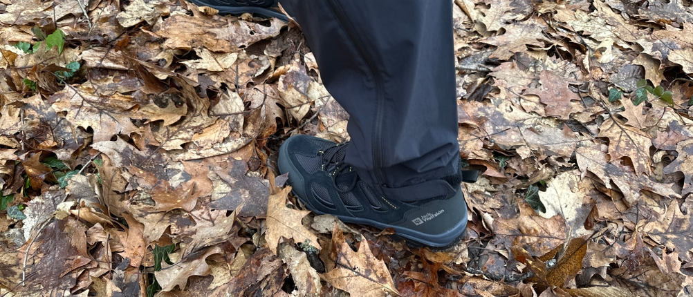 Die Rückansicht einer Vaude Herren Farley Stretch T-Zip Hose III auf herbstlichem Waldboden. Die Hose fällt locker über die Wanderschuhe und schützt die Beine.
