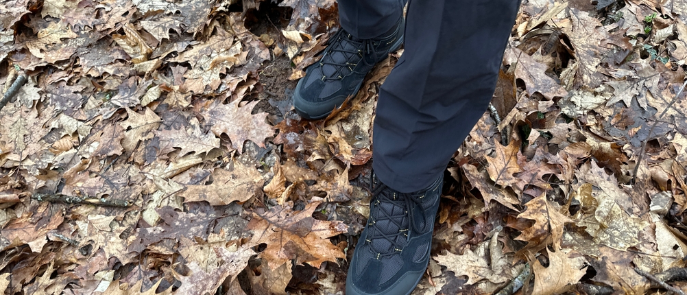Eine Person steht im Wald mit der Vaude Herren Farley Stretch T-Zip Hose III und schwarzen Wanderschuhen auf feuchtem Laub. Die Hose reicht bis zum Schuh und schützt die Beine.
