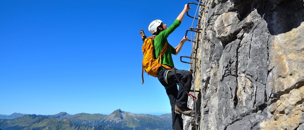 Ein Mann mit Rucksack und Helm erklimmt eine Eisenleiter im Klettersteig. Er trägt eine schwarze Wanderhose und ist gesichert.