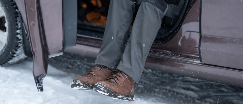 Eine Person sitzt in einem Auto mit geöffneter Tür und trägt eine zweifarbige Wanderhose. Draußen liegt Schnee, die Schuhe sind leicht verschneit.