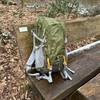 Der Mountaintop-TX5832lanpeicheng-Wanderrucksack auf einer Holzbank im Wald. Der grüne Rucksack steht aufrecht mit sichtbaren Riemen und Netzfächern.