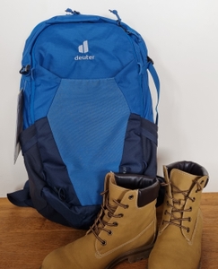 Der blaue deuter Wanderrucksack steht aufrecht neben einem Paar brauner Wanderstiefel. Beide befinden sich auf einem Holzboden vor einer weißen Wand.