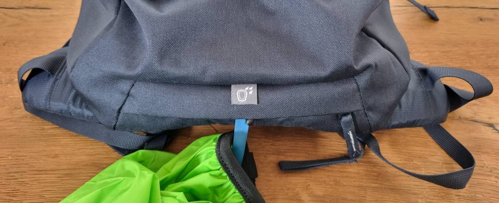 Eine grüne Regenhülle wird aus dem unteren Fach eines dunklen Wanderrucksacks mit Symbol herausgezogen. Der Rucksack liegt auf einem Holzboden.