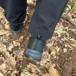 Rückansicht des Salewa Ms Alp Trainer Mid beim Gehen auf Waldboden. Die gelbe Vibram-Logo-Plakette ist sichtbar.