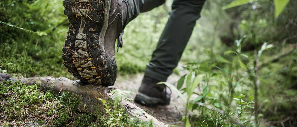 Ein Wanderer tritt mit Wanderschuhen auf nassem, glattem Untergrund im Wald. Die Schuhsohle ist mit Erde und Nadeln bedeckt.