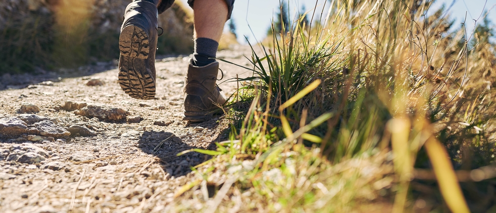 Ein Wanderer geht zügig über einen trockenen Schotterweg mit niedrigem Gras am Rand. Der Fokus liegt auf den Sohlen der Wanderschuhe.