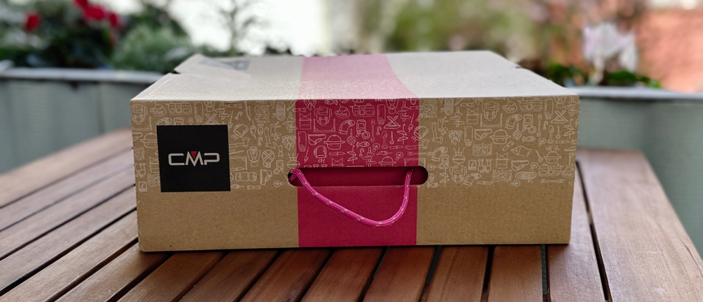CMP-Schuhkarton mit Tragegriff auf einem Holztisch, im Hintergrund unscharfe Blüten und Häuserfassaden.