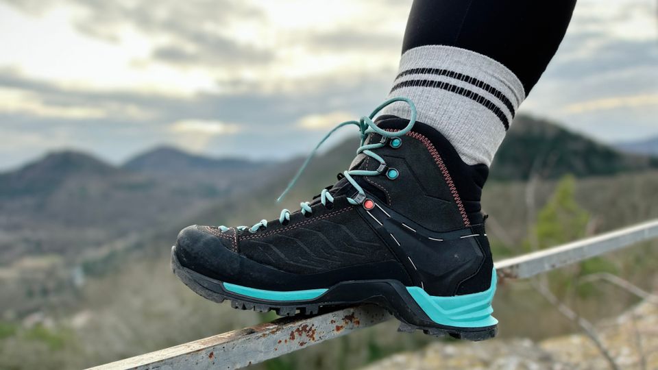 Zu sehen ist ein Bein-Ausschnitt einer Person, die den Fuß mit einem Wanderschuh auf ein Geländer stellt. Im Hintergrund sind Wald und Hügel zu sehen.
