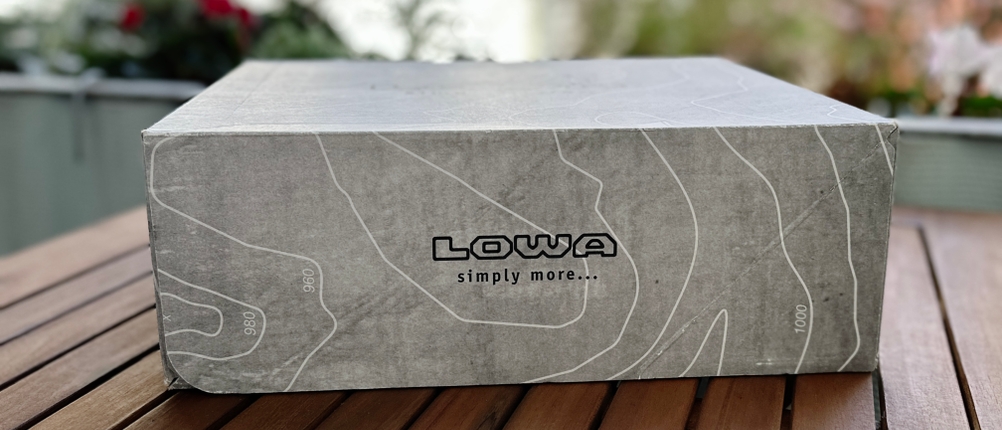 Lowa-Schuhkarton mit Höhenlinien-Design auf Holztisch. Der Slogan „simply more…“ ist aufgedruckt.