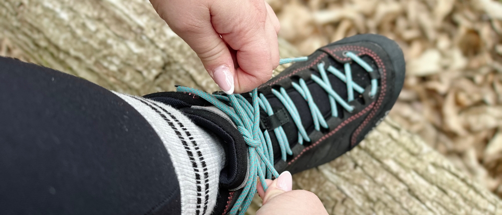 Zwei Hände binden die Schnürsenkel des Salewa MTN Trainer Mid GTX über einem gefällten Baumstamm.