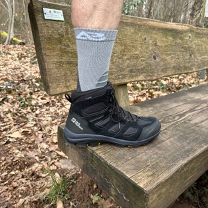 Eine Person testet den Sitz der Alpen Bears Premium Wandersocken, während der Fuß in einem Wanderschuh steckt. Die Szene findet auf einer Holzbank im Wald statt.