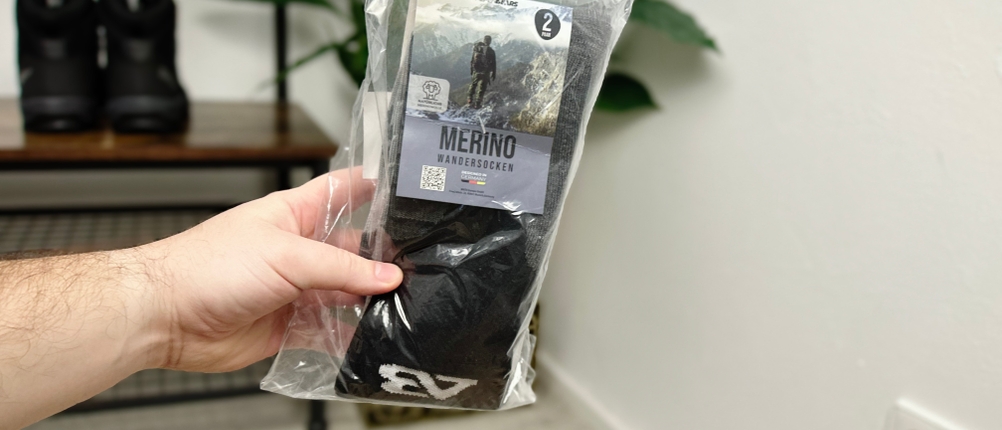 Eine Hand hält die originalverpackten Alpen Bears Premium Merino Wandersocken in einer durchsichtigen Plastikverpackung. Im Hintergrund stehen Wanderschuhe auf einer Bank in einem Raum mit einer Pflanze.