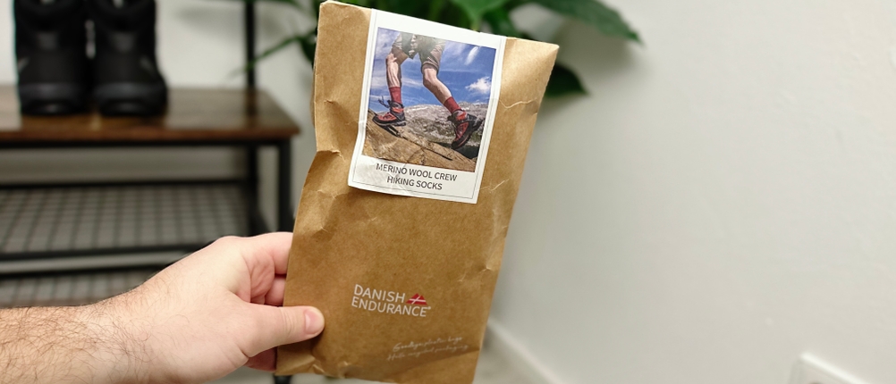 Eine Hand hält die originalverpackten Danish Endurance Merino Wandersocken in einer braunen Papierverpackung. Im Hintergrund stehen Wanderschuhe auf einer Bank.