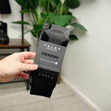 Eine Hand hält die ausgepackten Falke TK2 Wandersocken mit der Original-Kartonverpackung. Im Hintergrund sind Wanderschuhe und eine Pflanze zu sehen.