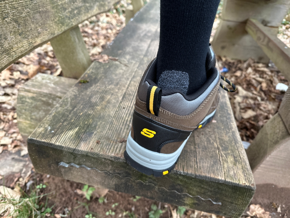 Ein Nahaufnahme-Bild zeigt einen Wanderstiefel mit einem Falke TK2 Wandersocken, der eine gepolsterte Ferse besitzt. Die Szene spielt sich auf einer Holzbank im Wald ab.