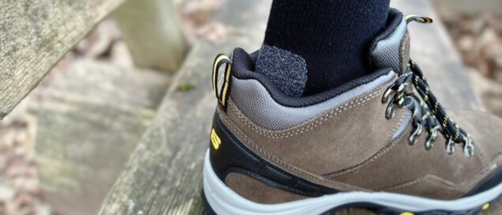 Ein Wanderschuh mit angezogenem Falke TK2 Wandersocken, wobei die gepolsterte Ferse der Socke hervorgehoben ist. Die Aufnahme wurde im Freien auf einer Holzbank gemacht.