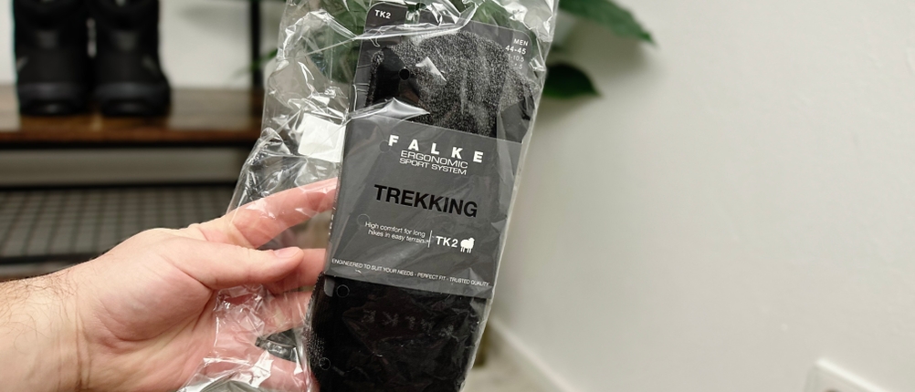 ALT-Text: Eine Hand hält die originalverpackten Falke TK2 Wandersocken in transparenter Plastikverpackung. Im Hintergrund stehen Wanderschuhe auf einer Bank in einem Raum mit einer Pflanze.