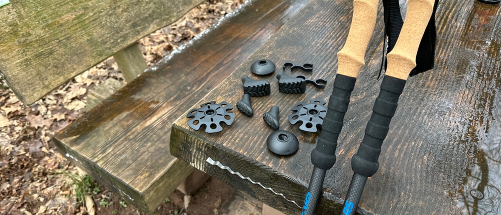 Die Alpen Bears Wanderstöcke liegen auf einer Holztischfläche, daneben verschiedene Aufsätze und Teller. Die Stöcke haben Korkgriffe und schwarze Schäfte.