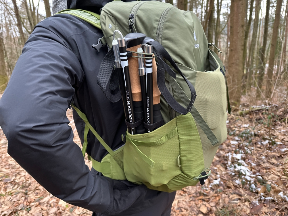 Ein Paar Alpin Loacker Wanderstöcke steckt seitlich im Außenfach eines grünen Wanderrucksacks. Die Szene spielt sich im Wald auf einem Laubweg ab.
