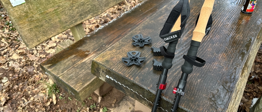 Zwei Alpin Loacker Wanderstöcke liegen auf einem Tisch, umgeben von Zubehör wie Gummipuffer und Tellern. Die Umgebung ist herbstlich-waldig.