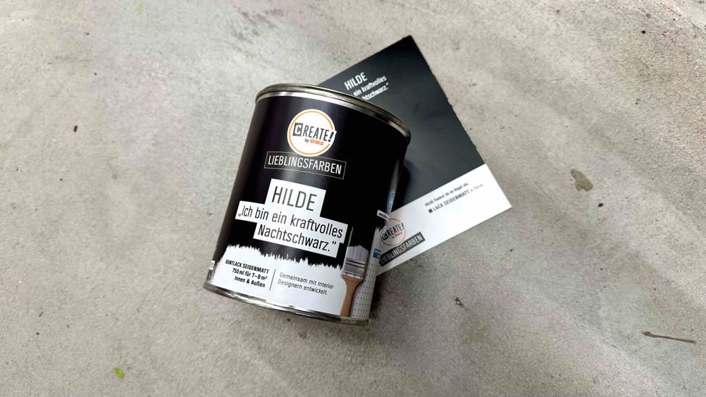 Auf der Farbkarte liegt eine Dose Wandfarbe Schwarz im Test auf einem Betonboden.