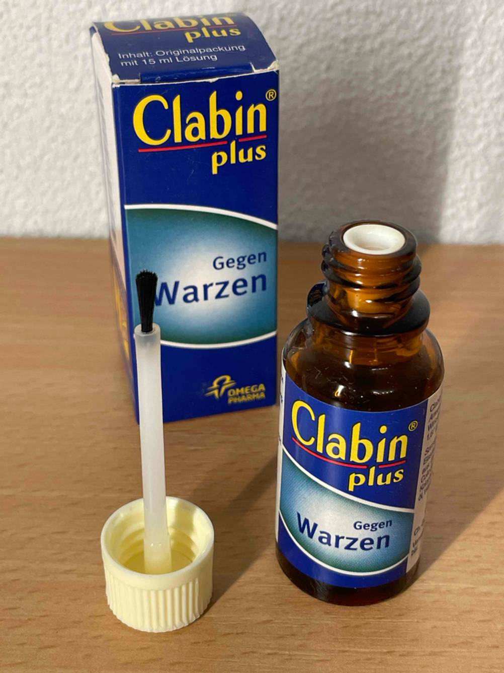 Die geöffnete Flasche des getesteten Warzenmittels steht neben dem Pinsel und vor der Originalverpackung auf einer Holzfläche.