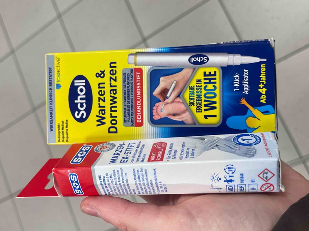 Im Test: Warzenstifte unterschiedlicher Hersteller liegen innerhalb ihrer gelb-blauen und rot-weißen Verpackung auf einer Handfläche.
