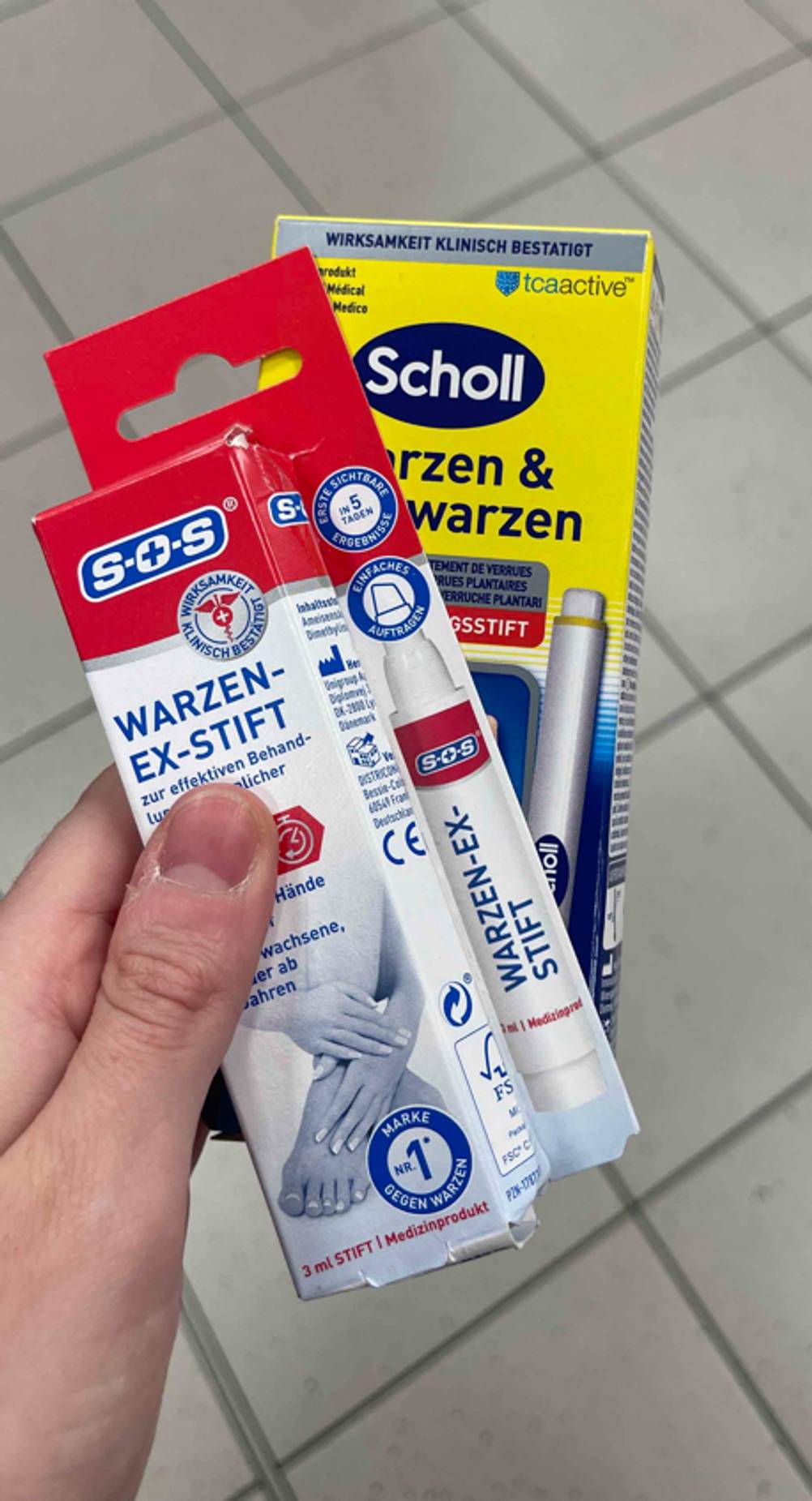 Warzenstifte im Test: Eine Hand hält zwei unterschiedliche Verpackungen über einem weißen Fliesenboden.