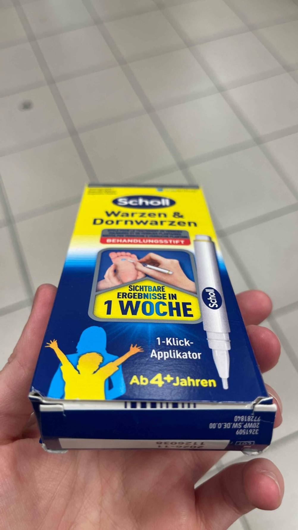 Warzenstift getestet: Eine Hand hält die gelb-blaue Verpackung der Firma Scholl über einem weißen Fliesenboden.