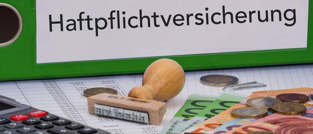 Haftpflichtversicherung-Test: Ein grüner Ordner mit der Aufschrift Haftpflichtversicherung liegt neben einem Taschenrechner, einem Stempel und etwas Geld.