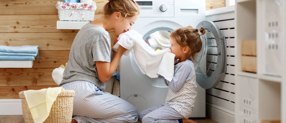 Eine Frau und ein Mädchen sitzen vor einer Waschmaschine und riechen an der frisch gewaschenen Wäsche.