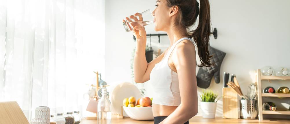 Eine junge Frau steht in der Küche und trinkt ein Glas Wasser.