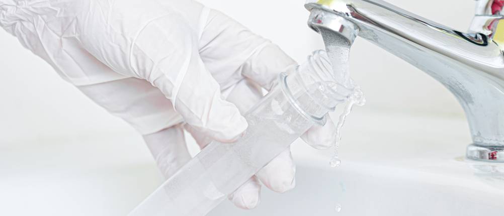Wassertest-Test: Eine Person mit Gummihandschuhen hält ein Reagenzglas unter den Strahl eines Wasserhahns.