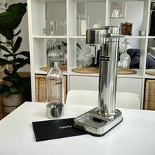 Der Aarke Carbonator 3 steht zusammen mit der Flasche und der Anleitung auf einem weißen Küchentisch. Das Gerät ist unbenutzt und sauber arrangiert.