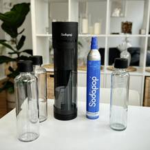 Der Wassersprudler Sodapop Logan steht auf einem Tisch, umgeben von drei Glasflaschen mit schwarzen Deckeln und einer blauen CO₂-Kartusche. Das Gerät und Zubehör sind ordentlich arrangiert, mit einem modernen Wohnraum im Hintergrund.