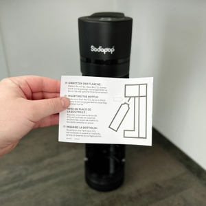 Eine Hand hält eine Anleitung vor den Sodapop Logan Wassersprudler. Das Dokument zeigt eine Skizze zum korrekten Einsetzen der Flasche.