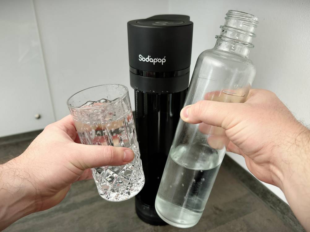 Sprudelwasser wird aus einer Glasflasche in ein Trinkglas gegossen. Der Sodapop Logan steht im Hintergrund.