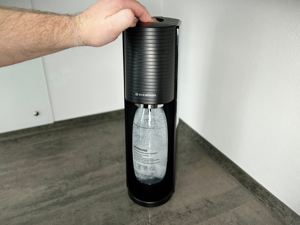 Ein Nutzer betätigt den Sprudelknopf des Sodastream Terra, während die Flasche im Gerät befestigt ist. Kohlensäure wird in das Wasser geleitet.