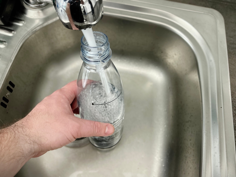 Eine Sodastream-Flasche wird mit Leitungswasser unter einem Wasserhahn befüllt. Das Wasser erreicht die Markierung für die maximale Füllmenge.