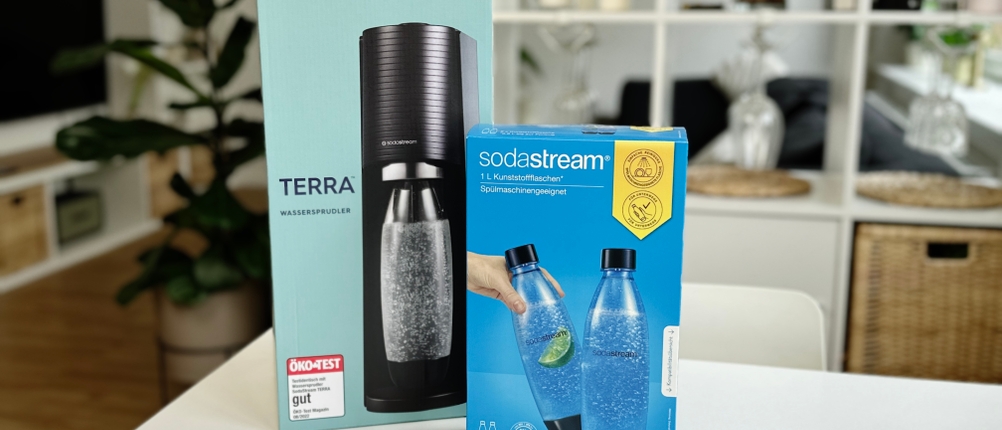  Der Sodastream Terra in seiner Originalverpackung steht auf einem Tisch. Daneben befindet sich eine Packung mit passenden Ersatzflaschen.