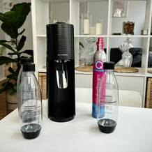 Der Sodastream Terra mit zwei Flaschen und einer CO₂-Kartusche auf einem Tisch. Das Gerät steht aufrecht, die Flaschen und der Zylinder sind daneben platziert.