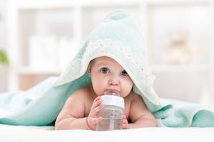Wassertest-Test: Ein Baby liegt auf einem Bett und schaut unter einer Decke hervor, während es eine Flasche Wasser in der Hand hält.