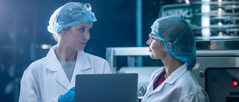 Zwei Frauen in Schutzkleidung stehen in einem Labor, während sie einen Laptop in den Händen halten.