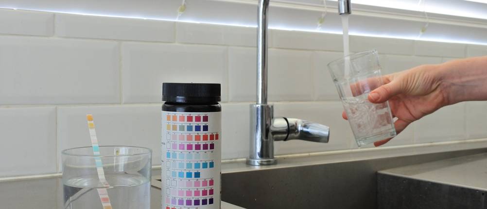 Ein Glas Wasser mit einem Testreifen steht neben einem Waschbecken, während eine Person sich an diesem gerade ein Glas Wasser abfüllt.