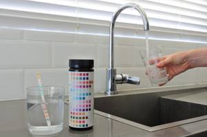 Wassertest-Test: Ein Glas Wasser mit einem Testreifen steht neben einem Waschbecken, während eine Person sich an diesem gerade ein Glas Wasser abfüllt.