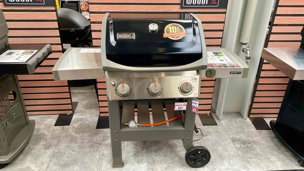 Ein hellgrauer Weber Stand-Gasgrill im Test inklusive orangem Gasanschluss-Schlauch befindet sich mit abgedeckter Grillfläche neben weiteren Grills in einem Geschäft.