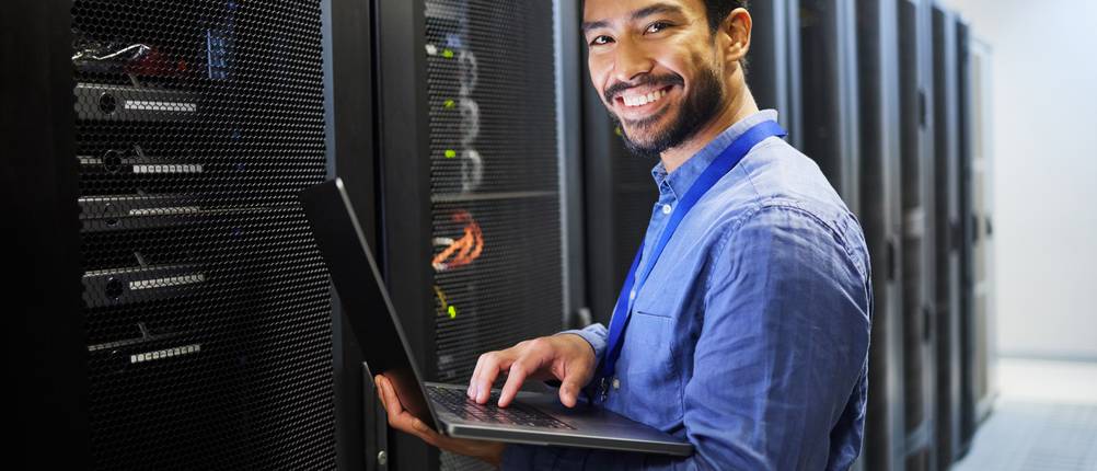 Webhosting getestet: Ein Mann im blauen Hemd steht mit einem Laptop im Arm vor einem Server-Schrank.