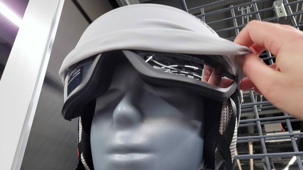 Hand hebt die hellgraue Schutzhaube eines bedeckten Visiers an, das zu einem grauen Skihelm mit Visier im Test gehört.