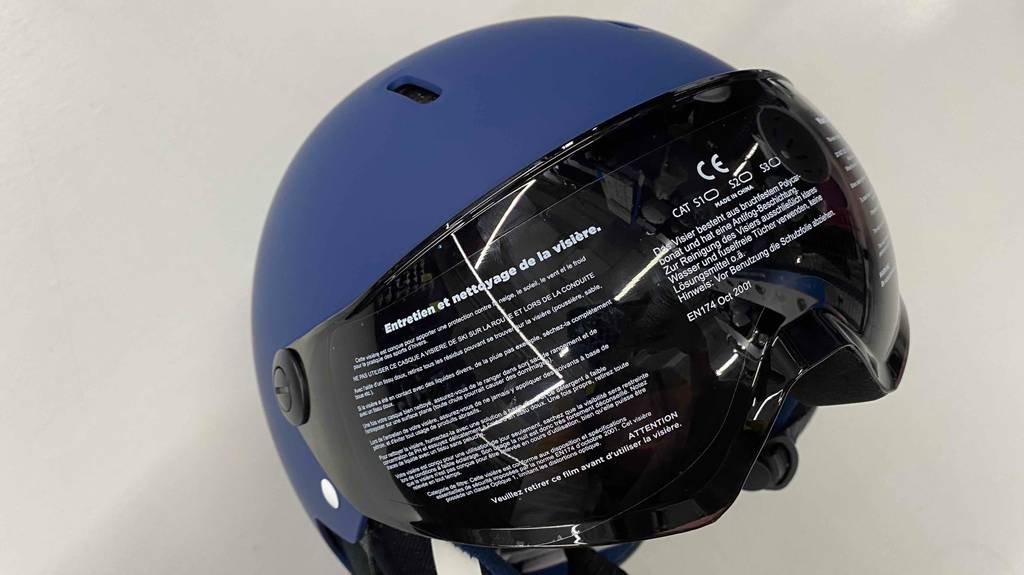 Vordere Detailansicht des abgeklebten, dunklen Visiers von einem blauen Skihelm mit Visier, der zum Test bereit auf einer hellen Fläche liegt.