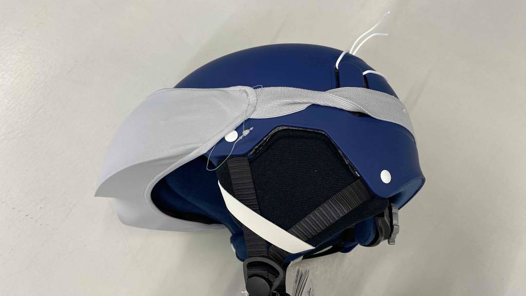 Seitliche Ansicht von einem Skihelm mit Visier im Test: blauer Skihelm mit hellem Visier-Schutzüberzug und schwarzen Befestigungsgurten liegt auf einem hellen Boden.
